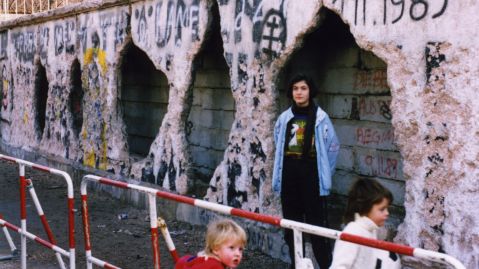 Die Berliner Mauer mit sichtbaren L&ouml;chern und Graffiti, eine Jugendliche und zwei Kinder davor.