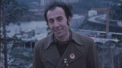 Iranischer Mann mit angestecktem Mikro an der Jacke lächelt in die Kamera vor einer Stadtansicht.