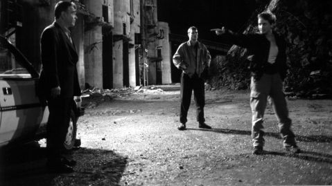 Three people on an industrial site at night, one pointing energetically, others standing quietly, concrete ruins in the background, scene looks tense.