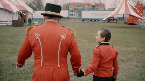 Ein Zirkusdirektor hält einen Jungen in ähnlichem Kostüm an der Hand und sie gehen gemeinsam in Richtung Zirkuszelt.