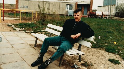 Ein junger Mann sitzt auf einer Bank. Seine Haare sind an den Seiten rasiert, er tr&auml;gt eine Bomberjacke, Jeans und Springerstiefel.