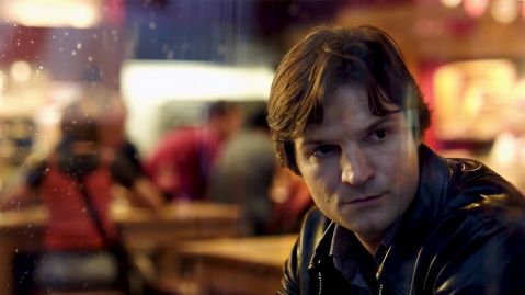 Ein Mann mit dunkelbraunem Haar sitzt in einem Restaurant, sein Gesicht ist ernst und konzentriert. Die Atmosphäre ist dunkel und still.