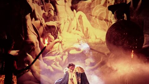 Ein Mann in königlicher Kleidung steht in einem Mantel vor einer Wand mit Soldatenfiguren und nackten Frauen.