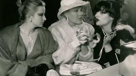 Drei elegant gekleidete Frauen sitzen auf einem Schwarzwei&szlig;foto nebeneinander und trinken Alkohol, eine spricht, zwei h&ouml;ren zu.