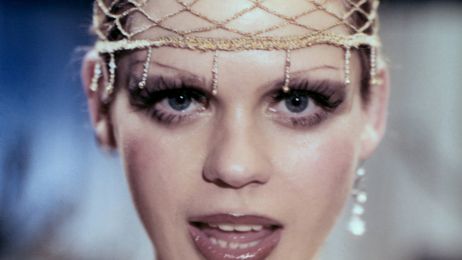 Eine stark geschminkte Frau blickt singend in die Kamera. A heavily made-up woman looks singing into the camera.