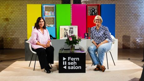 Die Schauspielerin und die Gastgeberin sitzen auf weißen Stühlen im Fernsehsalon, hinter ihnen bunte Balken.