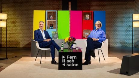 Zwei Personene sitzen auf einem Podium vor einem Hintergrund mit knalligen Farbfl&auml;chen.