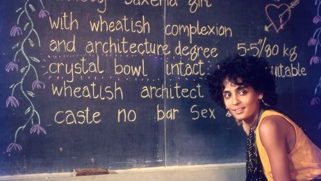 Frau sitzt vor einer Tafel, auf der viel Geschrieben steht und Blumen gemalt sind. Sie l&auml;chelt.