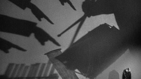 Ein Mann steht mit gebeugten Schultern in einem gro&szlig;en Raum, auf dem sich gro&szlig;e Schatten abtzeichnen.