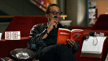 Ein schwarzer Mann mit Sonnenbrille sitz auf einer Couch. Er hat ein rotes Buch in der Hand und spricht auf ein Tonband. 