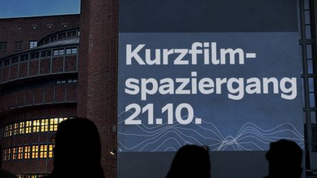 Menschen stehen draußen vor einer großen Leinwand mit dem Text „Kurzfilm-Spaziergang 21.10.“. Es dämmert und im Hintergrund ist das E-Werk mit beleuchteten Fenstern zu sehen. 