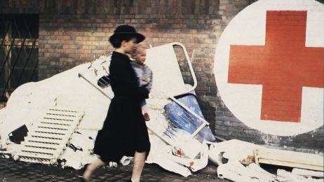 Woman with child walking past rubble and a Red Cross sign on a wall - a symbol of post-war and reconstruction.