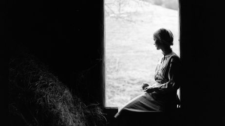 Eine Frau sitzt in einer Scheune am Fenster. Sie schaut nach drau&szlig;en und wirkt melancholisch. 