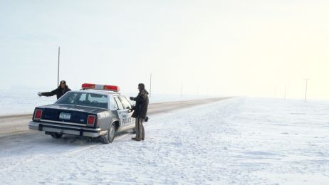 Ein altes Polizeiauto und zwei Polizisten stehen neben einer verlassenen Stra&szlig;e. Um sie herum ist nur Schnee und die lange Stra&szlig;e. 