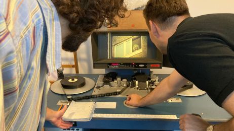 Zwei Männer von hinten schauen auf einen Bildschirm, auf dem ein Film mit einem Projektor abgespielt wird.