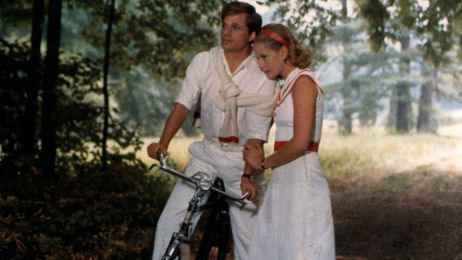 Zu sehen sind ein Mann auf dem Fahrrad und eine Frau an ihm gelehnt. Sie stehen im Wald und tragen wei&szlig;e Kleidung. 