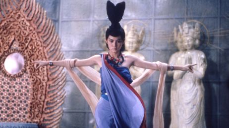 Eine Frau in blauem Gewand und mit au&szlig;ergew&ouml;hnlichem Kopfschmuck hat die Arme ausgebreitet und wird von hinten gehalten.