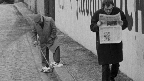Ein Mann l&auml;uft auf einem B&uuml;rgerstei und liest die Zeitung, w&auml;hrend ein anderer Mann in der N&auml;he mit seinem Spazierstock in einer fallengelassenen Zeitung stochert.