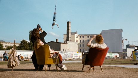 Two women are sitting on armchairs in a Berlin wasteland. They are looking at a bungee jumper hanging from a rope in the sky.