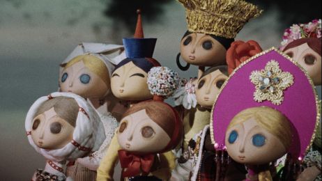 Szenenfoto aus einem Puppentrickfilm: Eine Gruppe von jungen Frauen in folkloristischer Kleidung blickt nach links auf ein Ereignis au&szlig;erhalb des Bildraums.