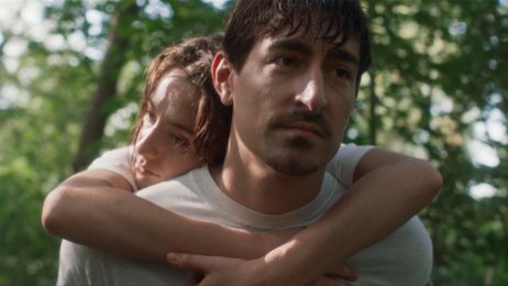 Eine junge Frau hängt schlapp auf den Schultern eines Mannes mit dunklen Haaren und Schnurrbart, der nach vorne schaut.