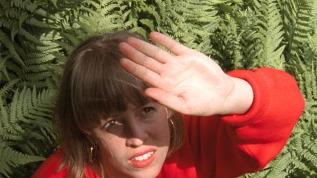 Eine Frau mit Pony und rotem Pullover steht vor Farn und hält sich schützend die Hand vor die Augen, um nicht von der Sonne geblendet zu werden.