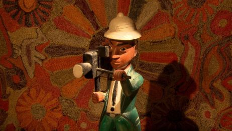 ChatGPT:  Eine Holzfigur mit einer kleinen Holzkamera steht vor einem teppichartigen Hintergrund mit Blumen.