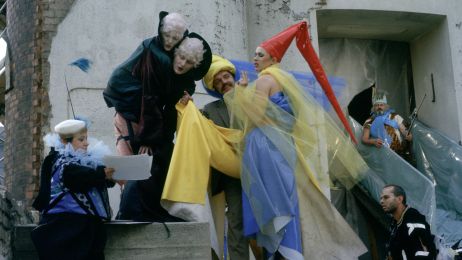 Mehrere Personen in bunten Kostümen stehen vor einem Gebäude und lesen etwas auf einem Blatt.