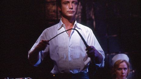 Ein Mann steht mit einer Peitsche im Raum, hinter ihm sitzt eine Frau.