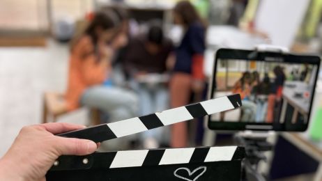 Im Vordergrund eine Hand, die eine Filmklappe h&auml;lt, im Hintergrund (unscharf) ein Tablet und eine Gruppe von Kindern.