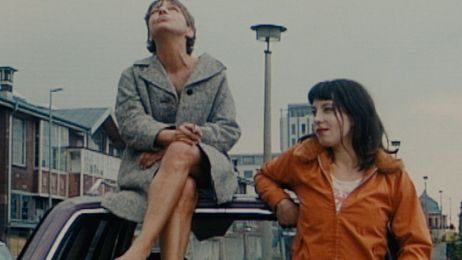 A barefoot woman sits on the roof of a car, next to her a younger woman leans and smiles.