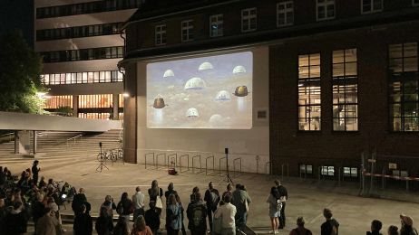 Es ist Abend und eine Menschengruppe steht vor zwei Häusern, ein Hochhaus im Hintergrund davor ein kleines älteres Haus auf dessen helle Außenwand ein Filmbild projiziert wird. 
