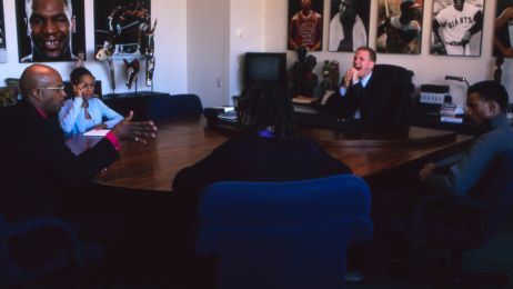 Eine Gruppe von Schwarzen Menschen und ein wei&szlig;er Mann sitzen um einen B&uuml;rotisch. An den W&auml;nden h&auml;ngen Fotos von Sportlegenden.