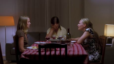 Three women are sitting at a table. The middle one holds her hands to her face while the other two look angrily at each other. The atmosphere seems tense.