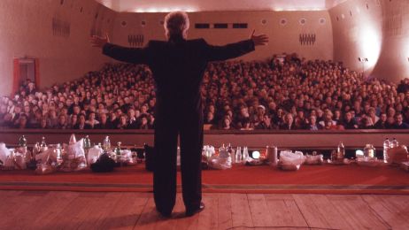 Ein Mann steht mit ausgebreiteten Armen auf der B&uuml;hne, der Publikumssaal ist voller Leute.