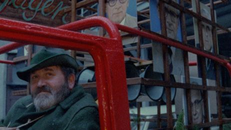 Dein Mann mit gr&uuml;nem Hut und Bart sitzt in einem roten Bus, im Hintergrund ein Haus mit einem Portr&auml;t Erich Honeckers.