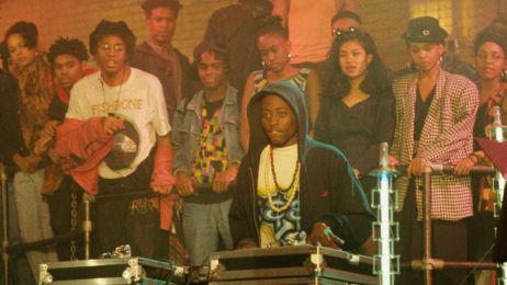 Ein junger Schwarzer Mann sitzt an DJ-Turntables, hinter ihm eine Gruppe von jungen Leuten, die ihm zuh&ouml;rt.