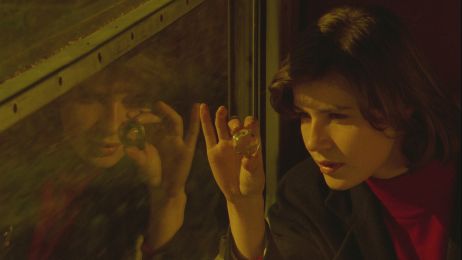 Eine junge Frau sitzt im Zug am Fenster und schat durch eine Glasmurmel. Sie spiegelt sich im Fenster.