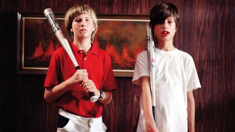 Zwei Jungen mit Baseballschl&auml;gern, einer in rotem, einer in wei&szlig;em T-Shirt stehen vor einer Holzwand. 