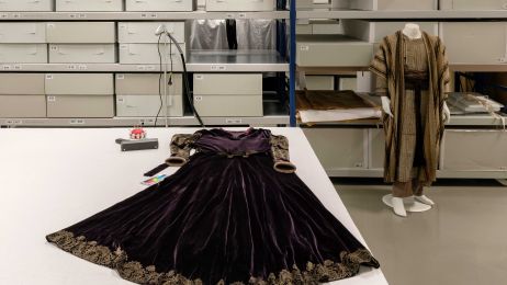 Ein dunkles Samtkleid mit Goldstickereien liegt ausgebreitet auf einem Tisch. Im Hintergrund Regale mit Archivk&auml;sten.