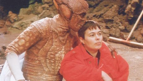 Costume designer Monika Bauert with an actor in a lizard costume on the film set. The actor hugs her and kisses her on the head.