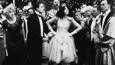 Die Schauspielerin Asta Nielsen steht in Kleid und Krone mit Armen in den H&uuml;ften in einer festlich gekleideten Gruppe und schaut herausfordernd zur Seite.