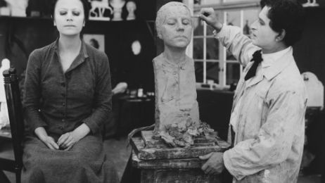 In einem Atelier sitzt eine Frau mit geschlossenen Augen einem Bildhauer Modell.