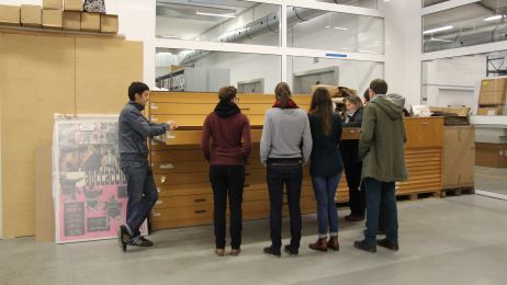 On the right, an archive employee explains posters from an open plan cabinet to a group of visitors.