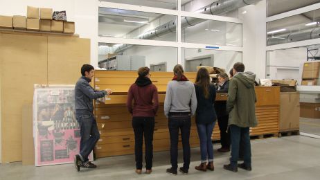 On the right, an archive employee explains posters from an open plan cabinet to a group of visitors.