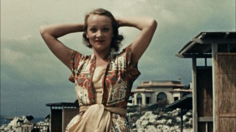 Marlene Dietrich steht in einem buntem Kleid vor Ruinen und H&auml;user. Sie hat die H&auml;nde hinter dem Kopf verschr&auml;nkt.