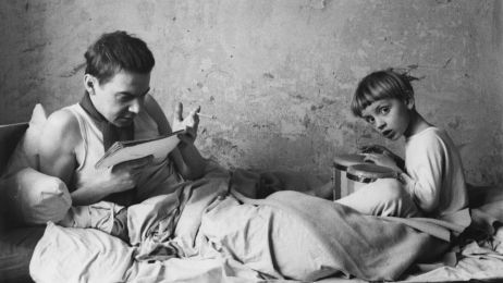 Ein Mann und ein Junge sitzen auf einem Bett. Der Junge spielt trommel, der Mann liest etwas und raucht.