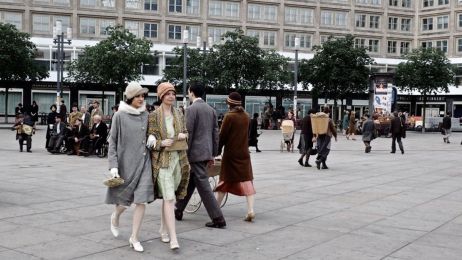 Menschen in 20er-Jahre-Kleidung flanieren &uuml;ber einen belebten Platz vor moderner Kulisse mit B&auml;umen und Schaufenstern.