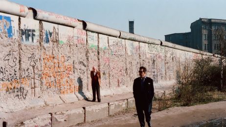 Mann im schwarzen Anzug und Sonnenbrille geht entlang der graffitibedeckten Berliner Mauer, w&auml;hrend ein anderer in einem leuchtend Anzug im Hintergrund steht.