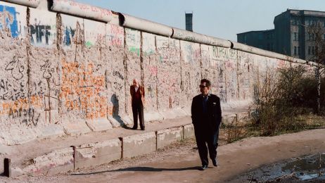 Mann im schwarzen Anzug und Sonnenbrille geht entlang der graffitibedeckten Berliner Mauer, w&auml;hrend ein anderer in einem leuchtend Anzug im Hintergrund steht.